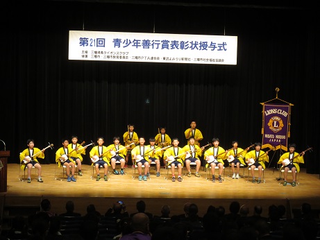 「第21回青少年善行賞表彰状授与式」と書かれた横断幕が掲げられた舞台の上で、黄色い法被を着た小学生たちが、前後2列に並んだ椅子に座って三味線を演奏し、その後ろで3人が太鼓を演奏している写真