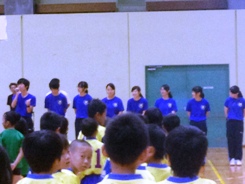 団体別に並んでいる子供たちの前で青色のシャツを着た8名の方々が横一列に並び、一番左に立っている方が話をしている様子の写真
