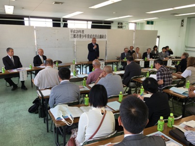 「平成28年度三郷市青少年育成市民会議総会」と書かれた横幕が後方の掲示ボードに貼られた室内で、長机の席に座っている参加者と、向かい合った席から立ち上がり挨拶を行っている市長の写真
