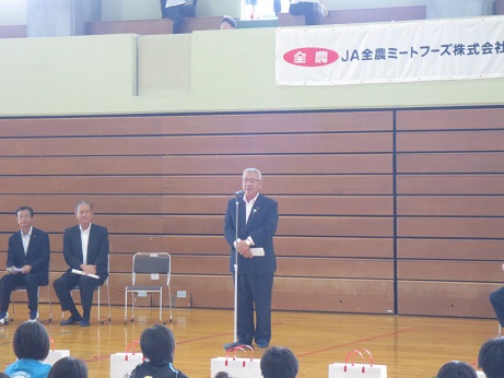 体育館内に設置されたマイクスタンドの前に立ち挨拶を行っている市長の話を、床に体育座りをして聞いている子供たちの後ろ姿の写真