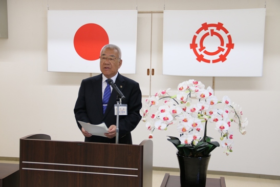 国旗と三郷市旗が吊り下げられている壁の前で、白い花が飾られた花台と並ぶ演台に立ち職員に訓示をする三郷市長の写真