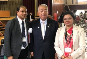 椅子の並んだ会場で、三郷市長が胸章を身に着けたネパール人男性とピンクのトップスの上に白のジャケットと黒のネックレスを着用したネパール人女性の間に立っている記念写真