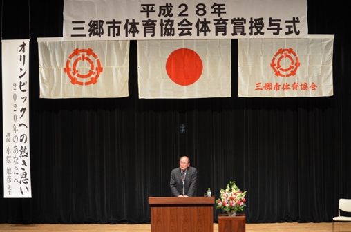 ステージ上部に「平成28年三郷市体育協会体育賞授与式」と書かれた横断幕が吊り下げられ、赤やピンク・白・緑の花で彩られた花台と並ぶ演台に立ち、「オリンピックへの熱き思い」のテーマについて講演を行う小原敏彦先生の写真