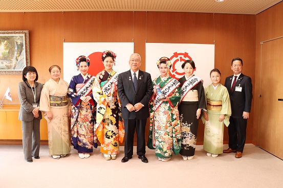 国旗と三郷市旗が飾られた室内で、市長を中央に6名の着物を着た女性とスーツを着た男性と女性が両端に立ち並んでいる記念写真