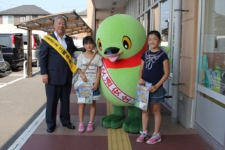 交通事故防止運動街頭キャンペーンのグッズを貰った2名の女の子がマスコットキャラクターかいちゃんの両側に立ち、左側に黄色のタスキをかけた市長と一緒に横一列に並んで立っている写真