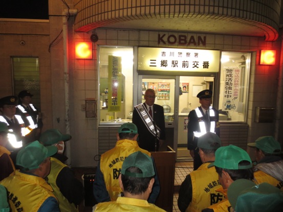 三郷駅前交番の入り口前に、緑の帽子や黄色のベストを着用した関係者が集まり、正面に立つ市長の方を向いている写真
