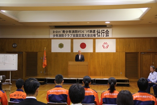「ヨーロッパ青少年消防オリンピック派遣・少年消防クラブ全国交流大会出場壮行会」と書かれた横断幕の下の檀上に市長が立ち話をしているのを椅子に座って聞いているクラブ員の後ろ姿の写真