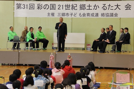 「第31回彩の国21世紀郷土かるた大会」と書かれた横断幕の下、可動式のステージの上でマイクを持った市長が話をしているのを体育館の床で体育座りをして聞いている子供たちの後ろ姿の写真