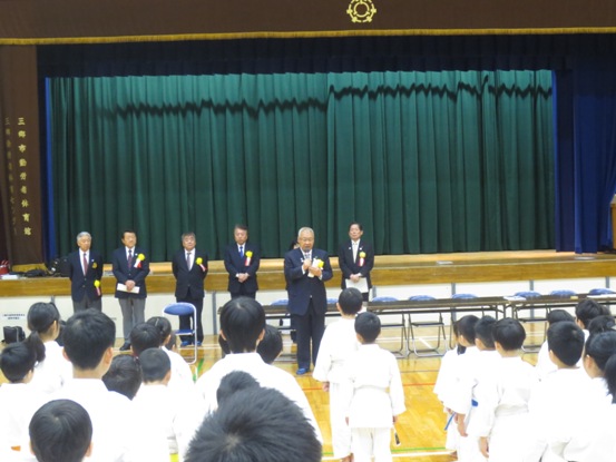 道着を着た子ども達が整列した体育館で、舞台下で黄色の胸章を着けた関係者たちが横一列に並んでいる前に市長が立って挨拶をしている写真