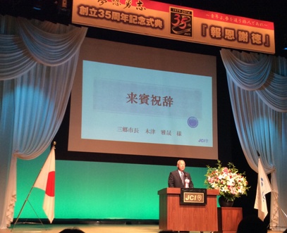 後方のスクリーンに「来賓祝辞」と書かれたステージ上の演壇で挨拶を行っている市長の写真