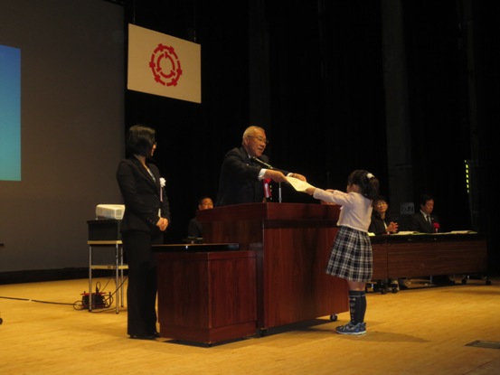 舞台上の演台から向かいに立つ女の子へ市長賞が贈呈されている様子の写真