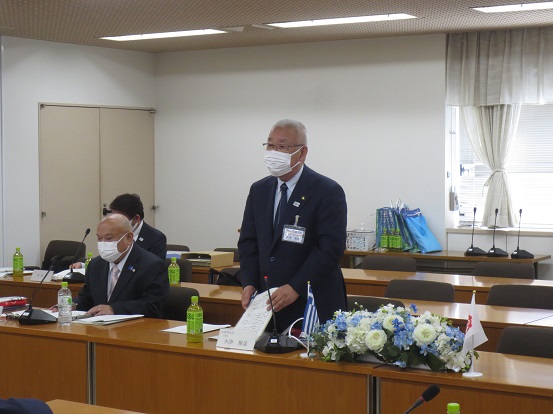 長机に水色や白の花と三郷市章とギリシャのミニ旗が飾られた席に立って挨拶している三郷市長と、席に座って聞いている関係者の方々の写真
