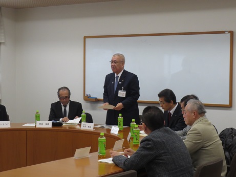 ホワイトボードの前の席で立ち挨拶の言葉を述べる三郷市長と、席札とお茶のペットボトルが置かれている席に座って話を聞く関係者の写真