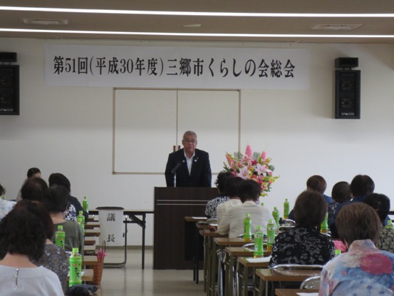 壁に「第51回(平成30年度」三郷市くらしの会総会」と書かれた紙が壁の上部に設置された部屋で、長机とともに用意されたパイプ椅子に着席している参加者たちと、正面に立って話をする市長の写真