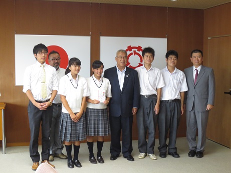 国旗と三郷市旗が飾られた室内で、市長を中央に4名の高校生と3名の先生方が横一列に並んでいる写真