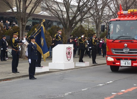 儀礼服を身につけた消防署員が歩道や歩道に置かれた演台などに立ち、道路を左前方へ向かって進む消防車を敬礼や旗を掲げて見送る様子の写真