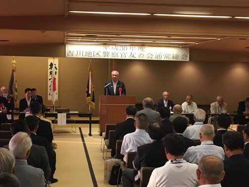 「平成30年度吉川地区警察官友の会通常総会」の紙が設置された和室で、椅子に座っている多くの関係者の正面に立って話をしている市長の写真
