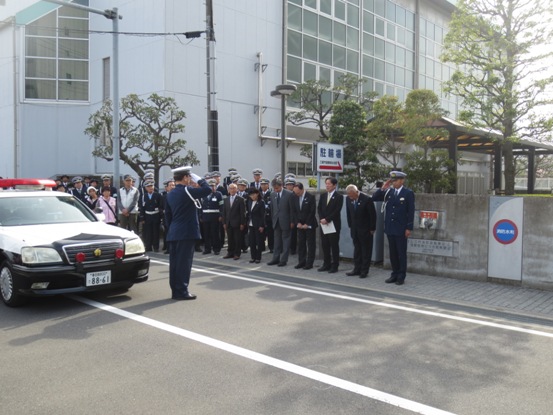 沿道に立つ多くの関係者たちと、道路に停まっているパトカーの前に立って敬礼をしている警察のかたの写真