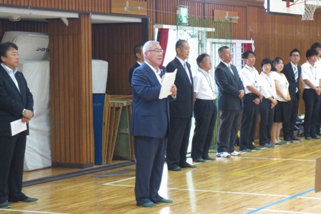 体育館の壁沿いに関係者が横一列に並び、手前に立つ市長が挨拶を行っている様子の写真