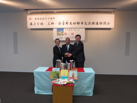 第30回「三郷・安曇野友好都市交流推進協議会」と書かれた横断幕が飾られた室内で、薄緑色のクロスがかけられたテーブルの上に絵本などの書籍が並んで置かれ、傍で市長を含め3名の町の代表者が握手をしている写真