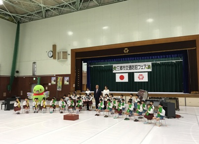 舞台前に置かれた打楽器やピアノの前で、扇形に2列に並んだ椅子に座った制服姿の幼稚園生たちと、その中央に立つ市長と白い服の女性、左側の園児の後ろで羽を広げて立つかいちゃんの記念撮影の様子を右斜めから写した写真