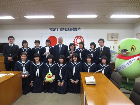 「平成29年度全国大会出場選手激励会」のポスターと国旗・三郷市旗が掛けられている壁の前で、それぞれかいちゃん・つぶちゃんのぬいぐるみ、トロフィーを持った女子生徒を含める女子チームのメンバーと引率の先生が、三郷市長とかいちゃんとともに並んで記念撮影をしている写真