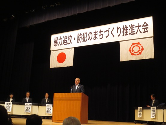 「暴力追放・防犯のまちづくり推進大会」と書かれた横断幕と国旗・三郷市旗が吊り下げられているステージ上で、演台に立ち挨拶の言葉を述べる三郷市長と、ステージ上に配置され役割名が書かれた用紙が貼り付けられている席に座り話を聞く関係者の写真