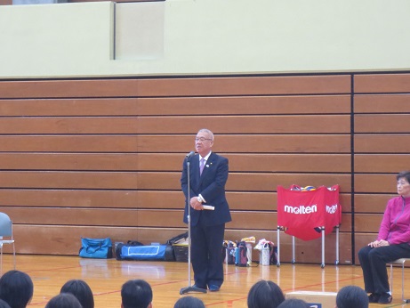 体育館でマイクに向かって立ち挨拶をする市長と、座って市長の方を向く子供たちの写真