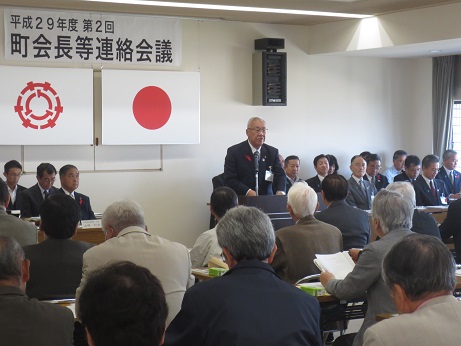 「平成29年度第2回町会長等連絡会議」と書かれた横断幕が貼り付けられ国旗と三郷市旗が吊り下げられている壁の前で、演台に立ち挨拶の言葉を述べる三郷市長と席に座って話を聞く関係者と町会長たちの写真