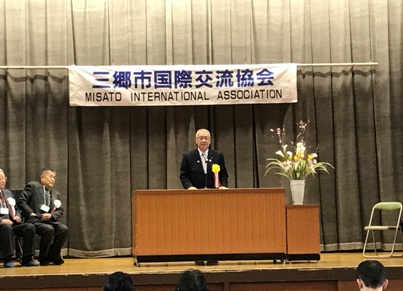 「三郷市国際交流協会」と書かれた横断幕が吊るしてあるステージ上で、ピンク・黄色・白の花々で彩られた花台と並ぶ演台に立ち挨拶の言葉を述べる三郷市長と、ステージ上に置かれたパイプ椅子に座り話を聞く胸章を付けた来賓者の写真