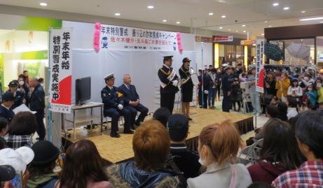 「年末特別警戒振り込め詐欺撲滅キャンペーン佐々木健介・北斗晶ご夫妻を迎えて」と書かれた横幕が貼られた小さなステージの左側に警察署長と市長が座り、佐々木健介さんと北斗晶さんがステージの上に立って話をしているのを大勢のお客さんが見に来ているキャンペーンの様子の写真