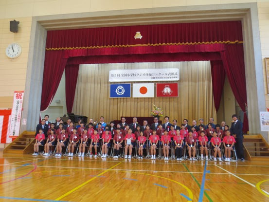 体育館の舞台の前に、メダルを肩から下げた小学生が横2列に並んでおり、後ろには関係者たちが一緒に並んで記念撮影をしている写真