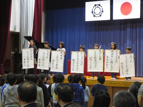 ステージ上に横一列に並び、新校舎にちなんだ俳句が書かれたポスター持ち披露する7名の子どもと、俳句を見ている子どもたちと関係者の写真