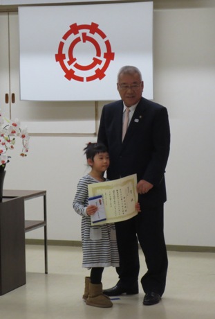 三郷市の旗が掲げられた壁を背に、表彰状を胸の前で正面に向けて持つ女の子と、その隣で手を携える市長を写した写真