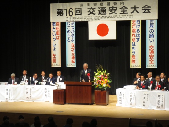 「第16回交通安全大会」と書かれた横断幕と国旗・交通安全に関する4つの標語が吊り下げられているステージ上で、赤・ピンク・黄色・白の豪華な花々で彩られた花台と並ぶ演台に立ち挨拶の言葉を述べる三郷市長と、ステージ上に配置された白いカバーが掛けられ席札が貼り付けられている席に座り話を聞く来賓者の写真