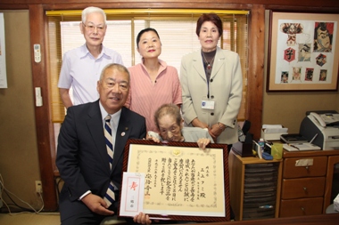 車いすに座って額縁に入った賞状を持った三上フミさんと市長が横並びに座り、後ろに3名の関係者が並んで立っている記念写真