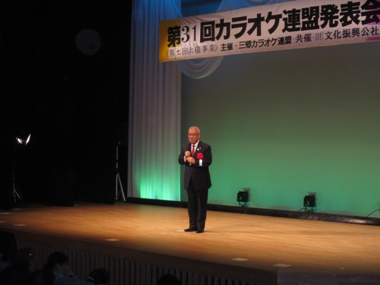 「第31回カラオケ連盟発表会」と書かれている横断幕が吊り下げられ、緑と青でライトアップされたステージ上で、赤い胸章を付けた三郷市長が話をしている写真