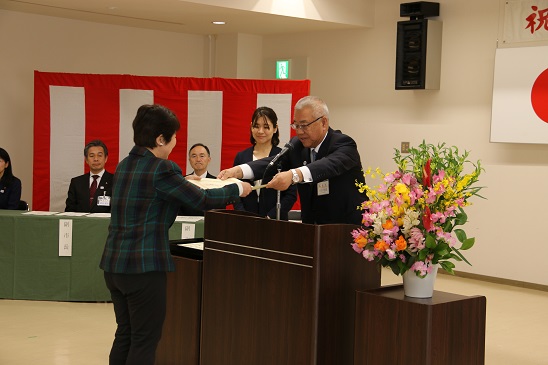 赤・ピンク・黄色・オレンジ・白の豪華な花々で彩られた花台と並ぶ演台に立ち表彰状を手渡す三郷市長と受け取る受賞者の女性、市長の横に立ち贈呈の様子を見守る介添人の女性の写真