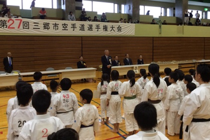 「第27回三郷市空手道選手権大会」と書かれた横断幕の下で挨拶を行っている市長の話を聞いている空手着を着た子供たちの後ろ姿の写真