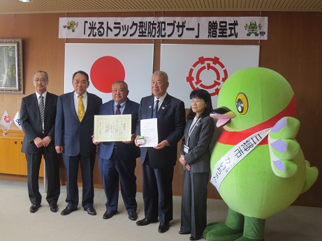 「光るトラック型防犯ブザー贈呈式」と書かれた横断幕が貼り付けられ国旗と三郷市旗が吊り下げられている壁の前で、黄緑色の防犯ブザーを持った三郷市長と埼玉県トラック協会吉川支部のメンバーが白いタスキをかけたかいちゃんとともに記念撮影をしている写真