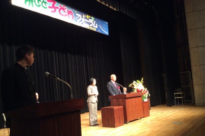 「みさと子どもフォーラム」と書かれた横断幕の下ステージの演壇で挨拶を行っている市長を左側に設置された演壇から見ている男子生徒の写真