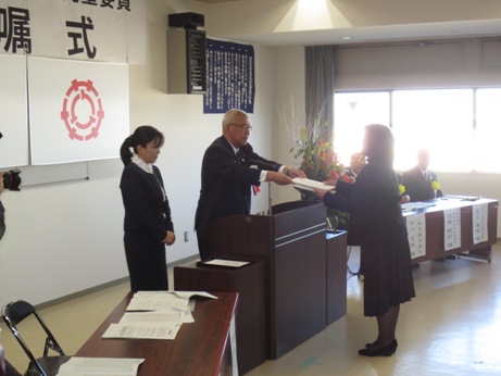 演台に立ち委嘱書を渡す市長と、それを受け取る女性、市長の横に控える女性職員、演台横の来賓席に座る男性たちを写した写真