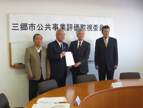 壁に設置されたホワイトボードの上半分いっぱいに「三郷市公共事業評価監視委員会」と印刷された紙が貼られ、ホワイトボードを背に立ち書類を胸の前で持った市長と、市長を挟んで立つ3人の委員が記念撮影をしている写真