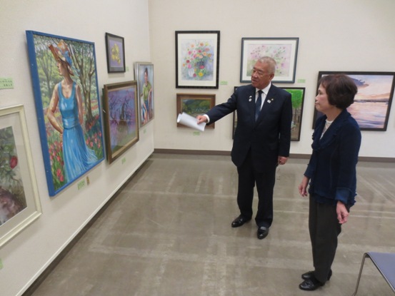 多くの美術作品が壁に展示されている部屋で、市長と女性が一つの作品の前に立って話をしている写真