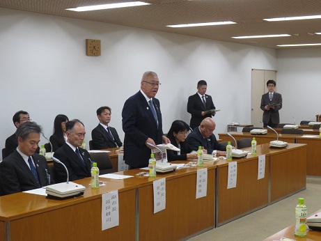 席を立ち北中ハンドボール部のメンバーへ激励の言葉を述べる三郷市長と、席札が貼られマイクとお茶のペットボトルが置かれている席に座り話を聞く関係者の写真