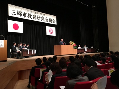 ステージの左右に設置された長机の席に座っている関係者や観客席に座っている方々が「平成28年度三郷市教育研究会総会」と書かれた横断幕の下の演壇で挨拶を行っている市長の話を聞いている写真