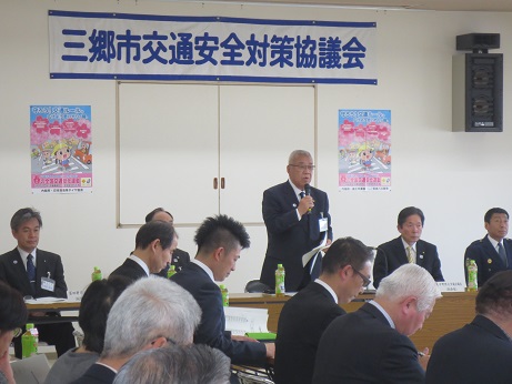 「三郷市交通安全対策協議会」と書かれた横断幕と桜の木と横断歩道を渡る子どもが描かれたパンフレットが貼り付けられている壁の前で、席を立ち挨拶の言葉を述べる三郷市長とペットボトルのお茶が入った席に座り話を聞く関係者の写真
