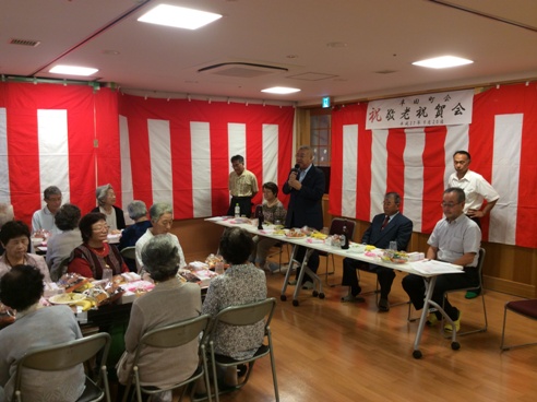 壁全体に紅白幕が貼られた会場で、マイクを持ち挨拶を行っている市長の話を着席して聞いている参加者の写真