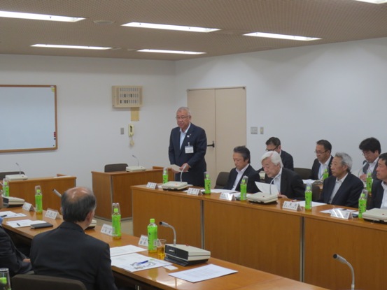 資料を手に持ち挨拶の言葉を述べる三郷市長と、席札とお茶のペットボトルが置かれた席に座り資料に目を通しながら話を聞く参加者の写真