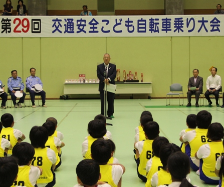 第29回交通安全こども自転車乗り大会と書かれた横断幕の手前に設置されたマイクの前に市長が立ち話をしているのを体育座りをして聞いている児童達の後ろ姿の写真